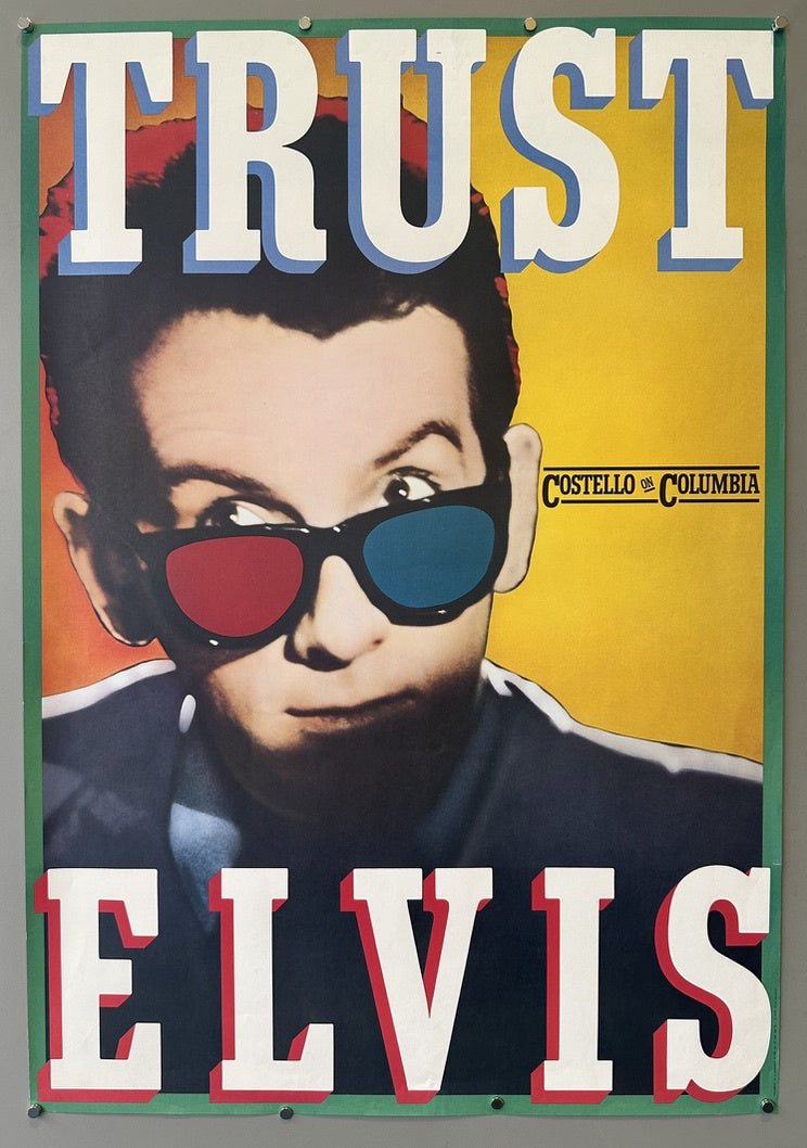 A promotional poster for Elvis Costello's 1981 album "Trust," featuring a close-up image of Elvis Costello wearing red and blue 3D glasses. The word "TRUST" is prominently displayed in large white letters with a blue outline at the top of the poster, and "ELVIS" is similarly displayed at the bottom.