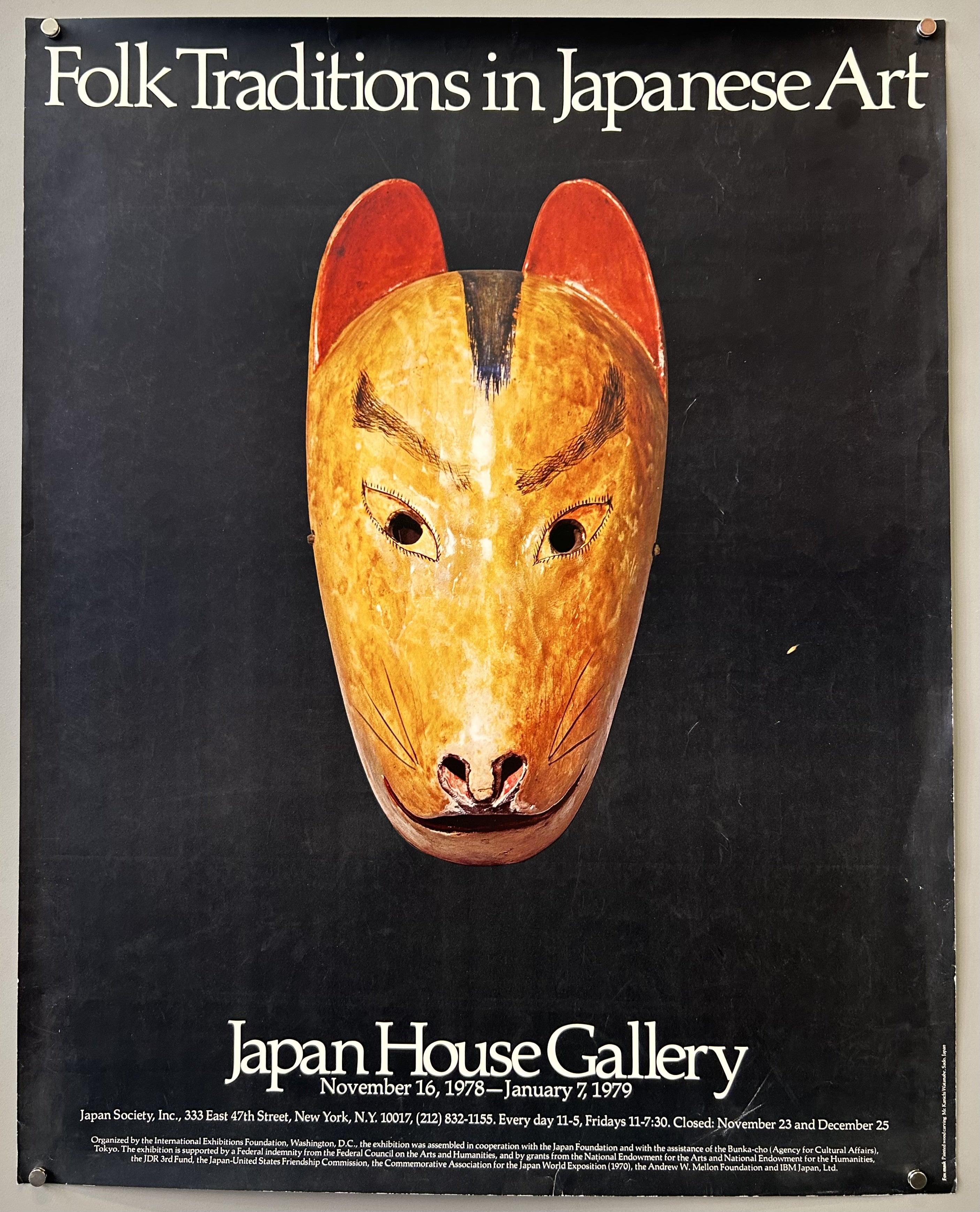 exhibition titled "Folk Traditions in Japanese Art" at the Japan House Gallery. The poster features a close-up, centered image of a wooden mask resembling a fox or dog, with red-tipped ears, against a black background. The text below the mask provides the exhibition dates (November 16, 1978 – January 7, 1979) and location details for the Japan Society in New York.