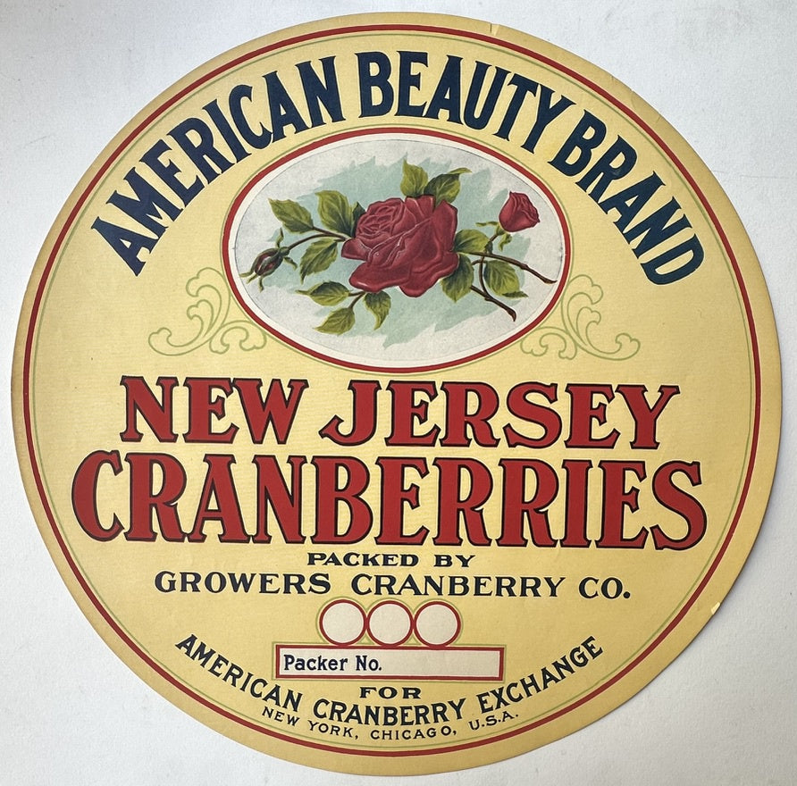 A round vintage-style label for "AMERICAN BEAUTY BRAND NEW JERSEY CRANBERRIES," featuring a central illustration of a red rose and rosebuds on a light background. Text below the illustration reads "PACKED BY GROWERS CRANBERRY CO."