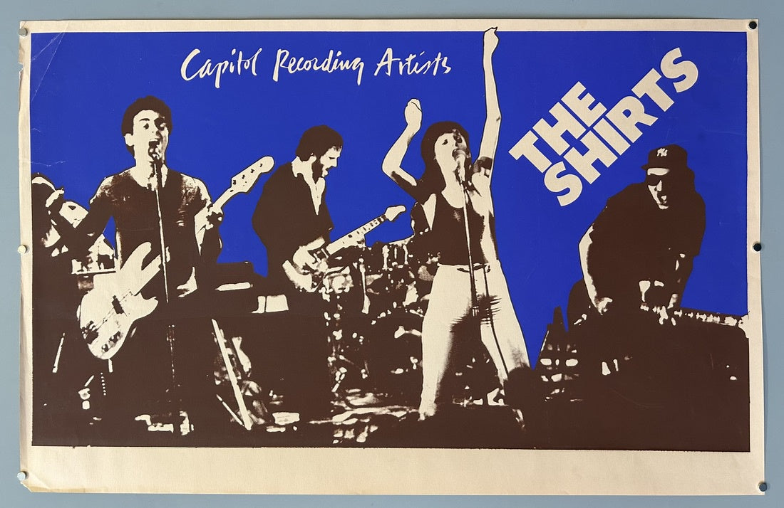 The poster features the band "The Shirts," identified as "Capitol Recording Artists," set against a striking blue background with brown/sepia-toned imagery. The design depicts four musicians performing on stage, with the band's name prominently displayed vertically on the right side of the poster.