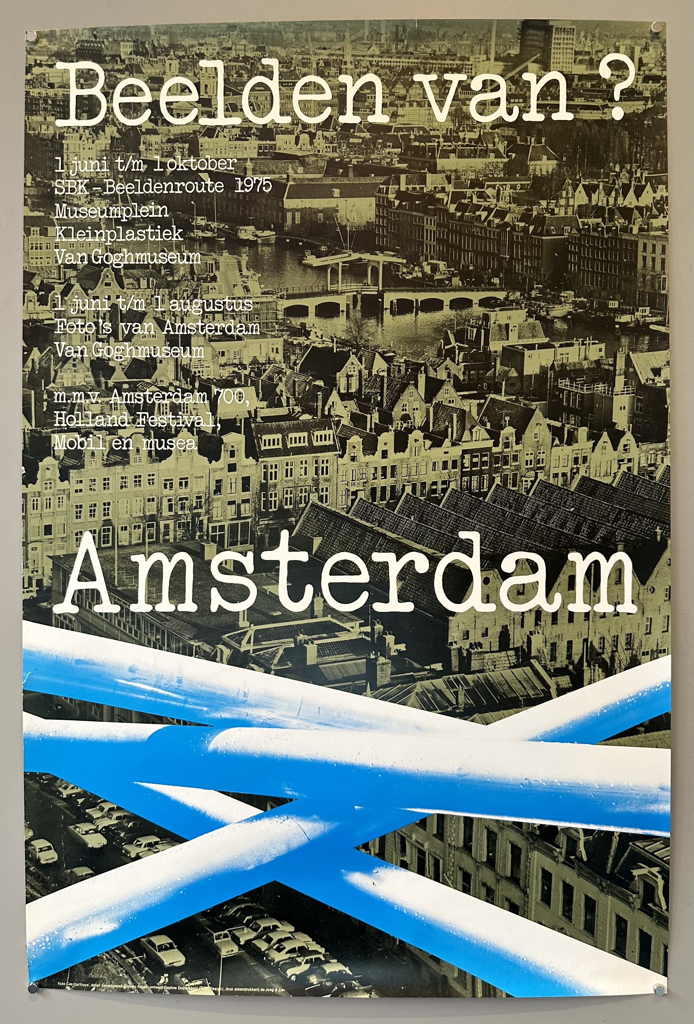 A vintage poster in Dutch for an exhibition in Amsterdam. The poster features a blue and white aerial view of Amsterdam with the word "Amsterdam" prominently displayed in the foreground.