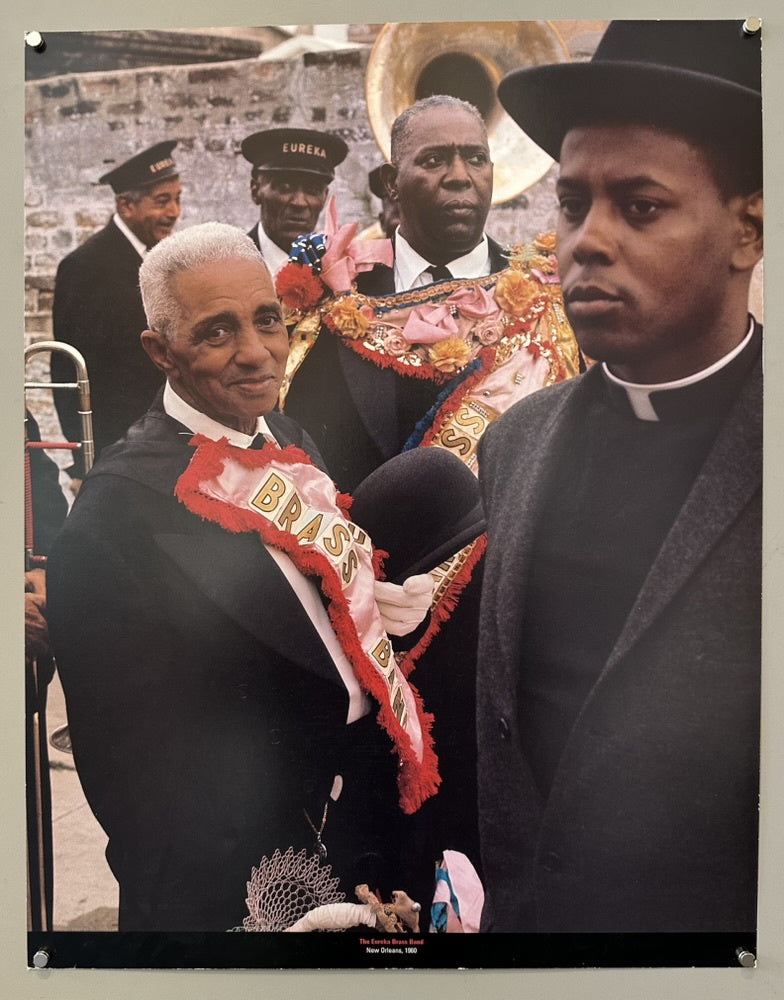 A color photograph depicting a group of African American men dressed in formal attire, likely a brass band, participating in a parade or procession. In the foreground, an older man with white hair and a sash reading "BRASS" smiles towards the left.