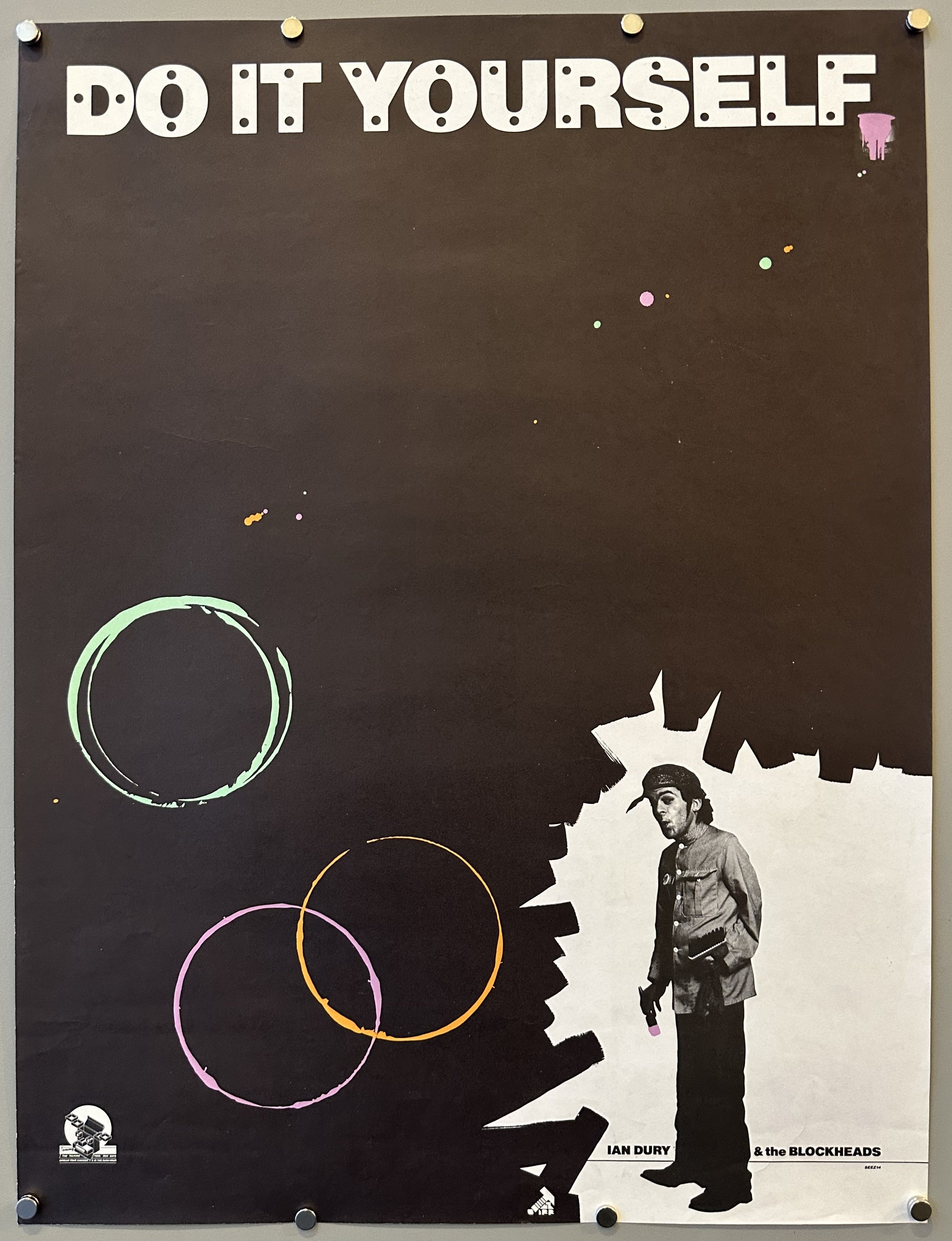 Black poster with pink, orange, and green splotches of color and Ian Dury's black and white figure on the bottom right-hand corner.