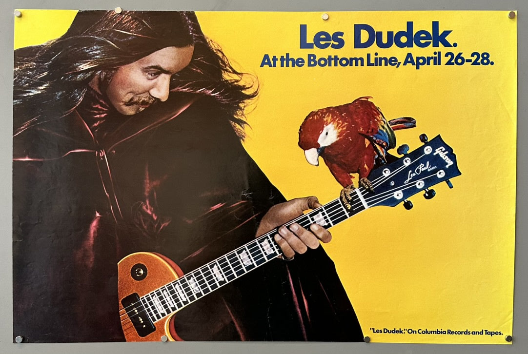 A concert poster for Les Dudek at The Bottom Line, April 26-28. The image features Les Dudek, a man with long dark hair and a mustache, wearing a dark cloak, holding a Gibson Les Paul guitar. A red macaw is perched on the headstock of the guitar. Below the image, text reads: "Les Dudek!" On Columbia Records and Tapes.