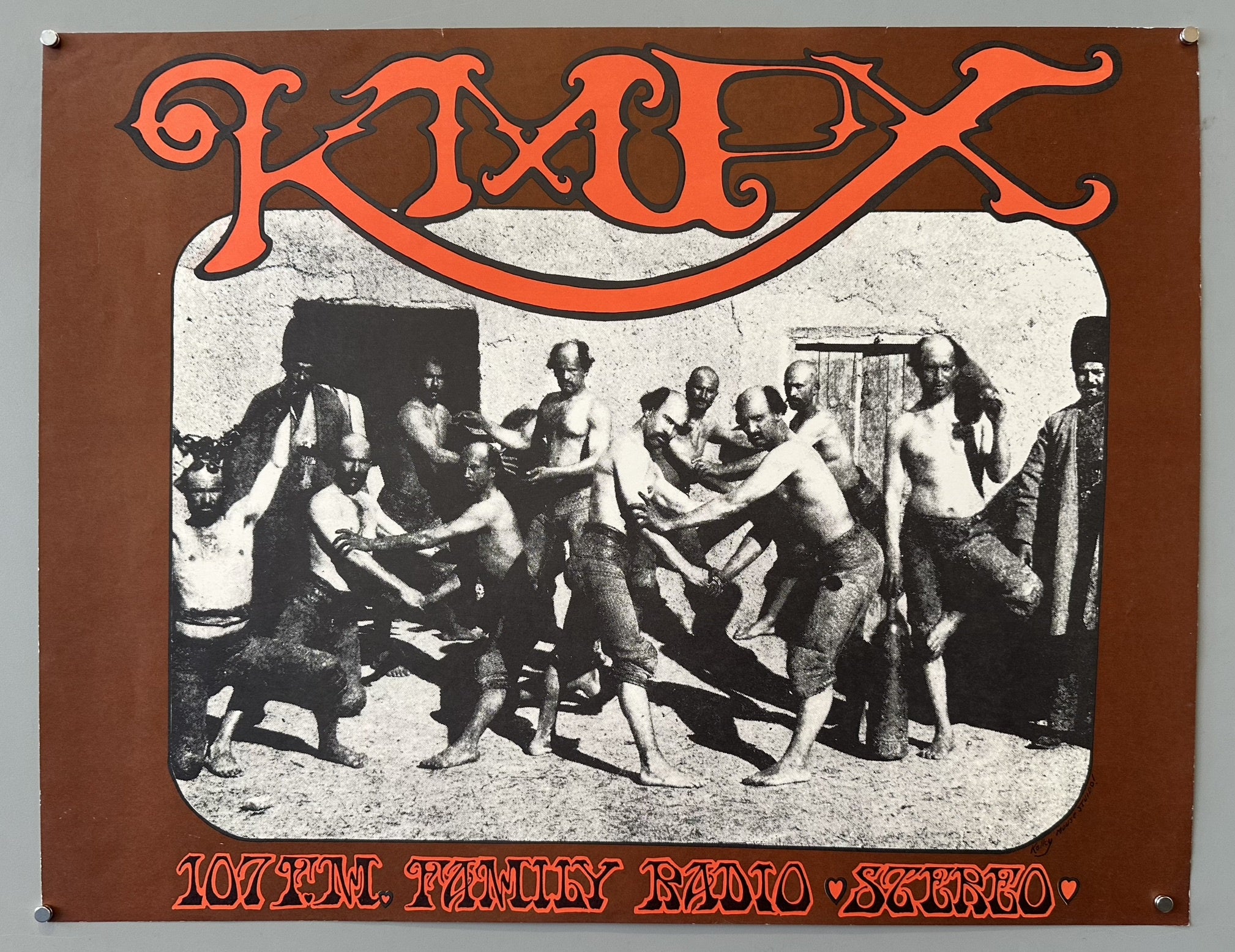 A brown KMPX 107 FM Family Radio Stereo poster featuring a black and white image of several men in various states of undress, engaged in what appears to be a physical struggle or game, set against a light-colored wall.