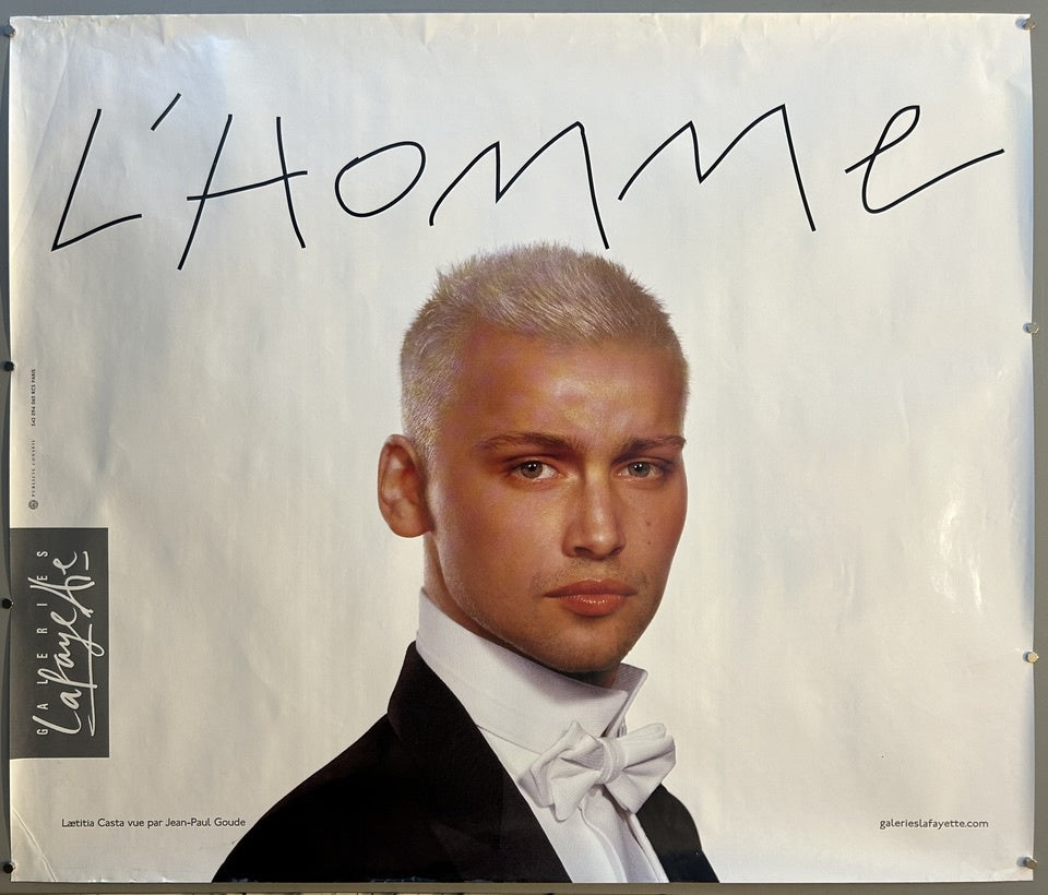 A promotional poster for Galeries Lafayette featuring a close-up portrait of a man with short, light-colored hair, wearing a tuxedo with a white bow tie. The title "L'Homme" is written in large, stylized script above his head.