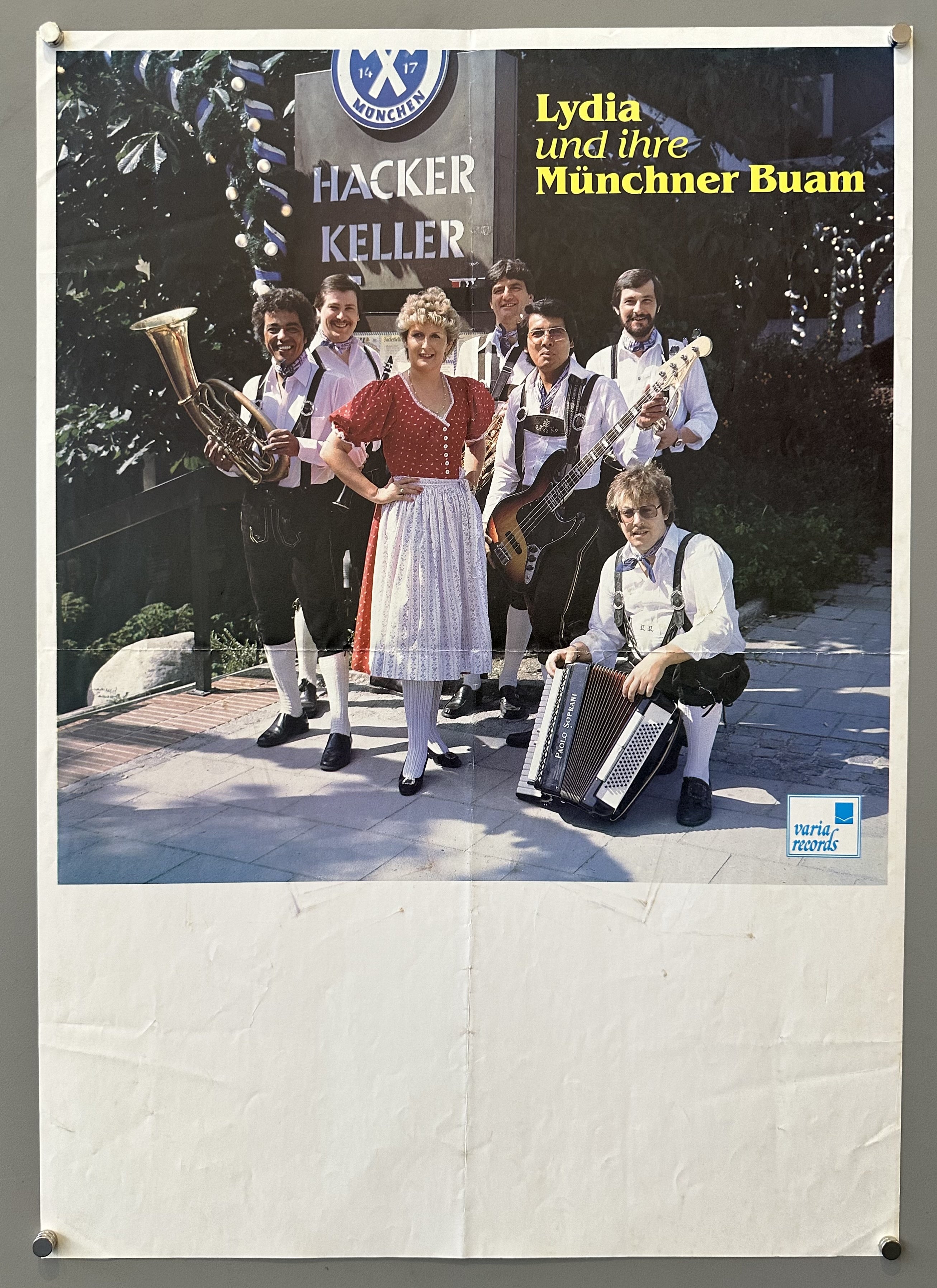 A vintage poster featuring a group of musicians in traditional Bavarian attire, including a woman in a dirndl dress, and men in lederhosen. They are posing outdoors, with a building featuring a sign that reads "HACKER KELLER"