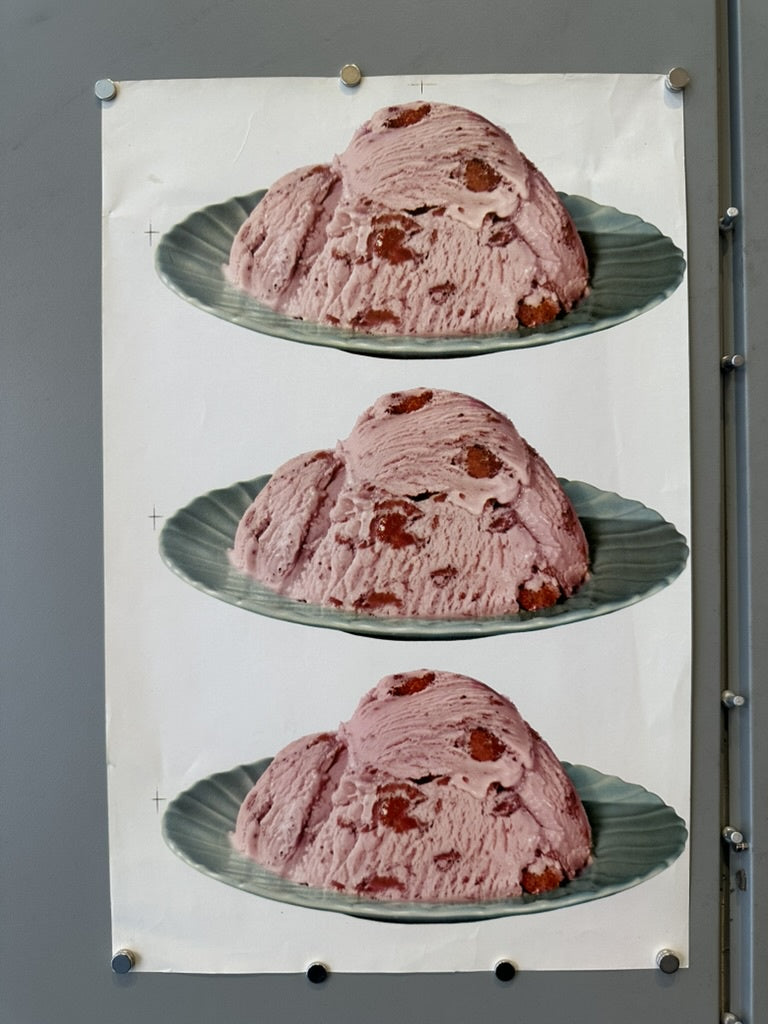 This image shows a poster with three identical scoops of strawberry ice cream on blue plates, pinned to a gray wall. The ice cream is pink with visible chunks of strawberries, and the plates have a scalloped edge.