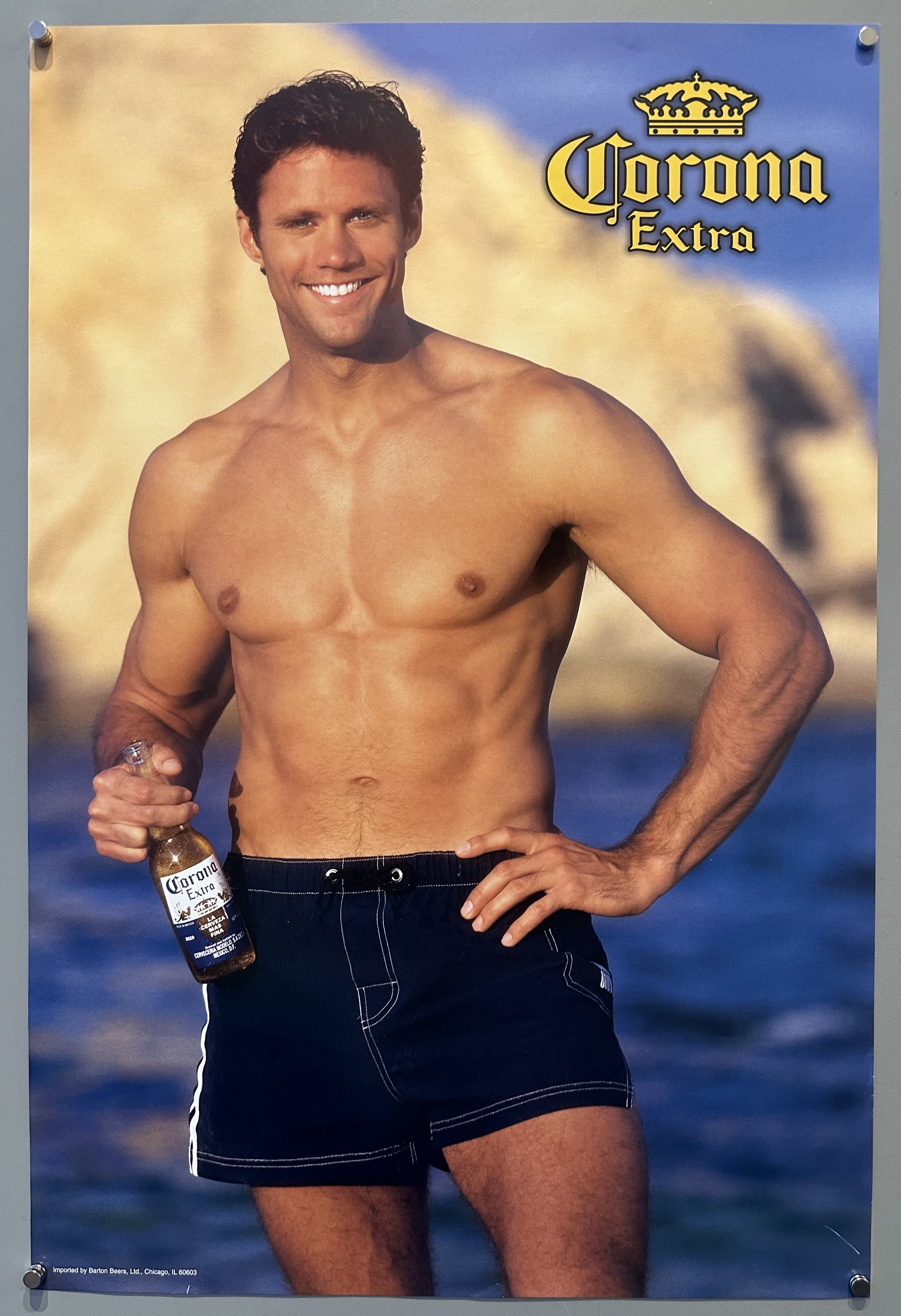 Shirtless man holds a Corona Extra while standing on a beach.
