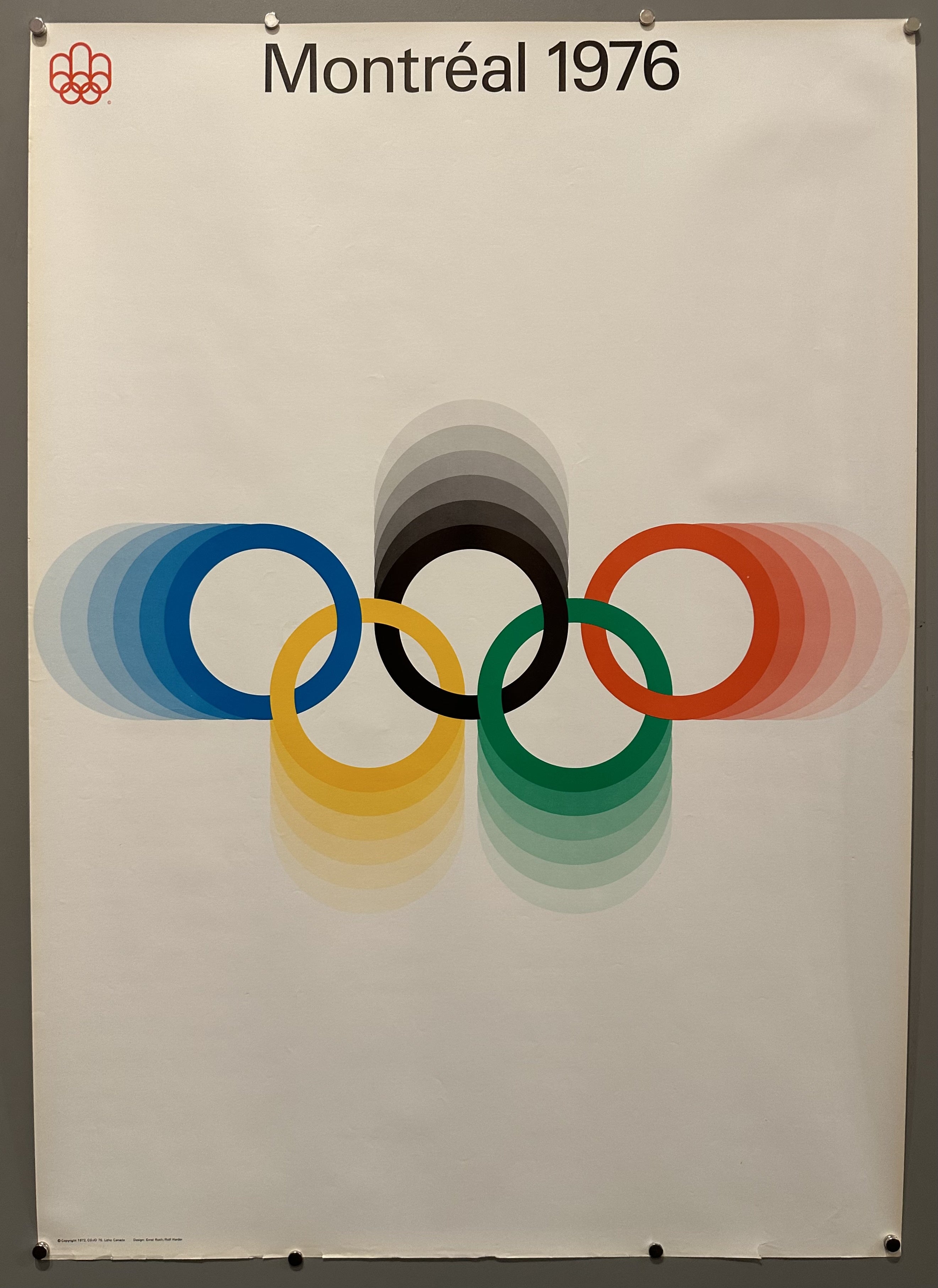A Montréal poster advertising the 1976 olympics going to take place in Canada. The Olympic logo of colorful rings is in the center with the colors gradually fading around it into a white background.