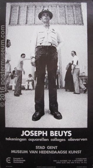 Black and white photograph of Joseph Beuys in front of a white building with other people in the background, staring at Beuys.