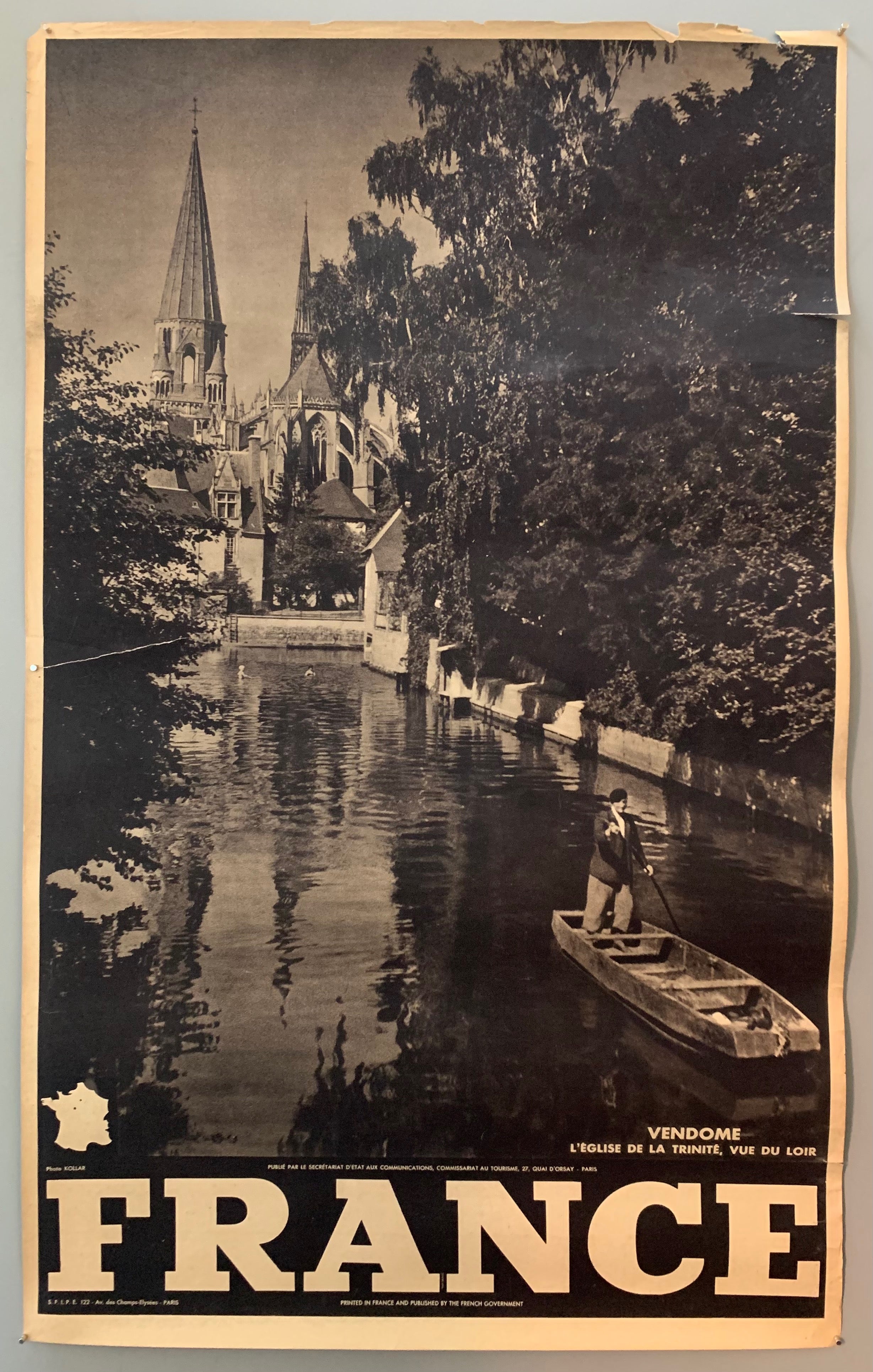 This poster is sepia toned with bold black-and-white writing at the bottom, as well as a tiny map of France. The photograph takes up the majority of the page.The photograph contains a view of a serene canal with a man boating down it. The river is framed by trees and in the background a gothic church is shown.