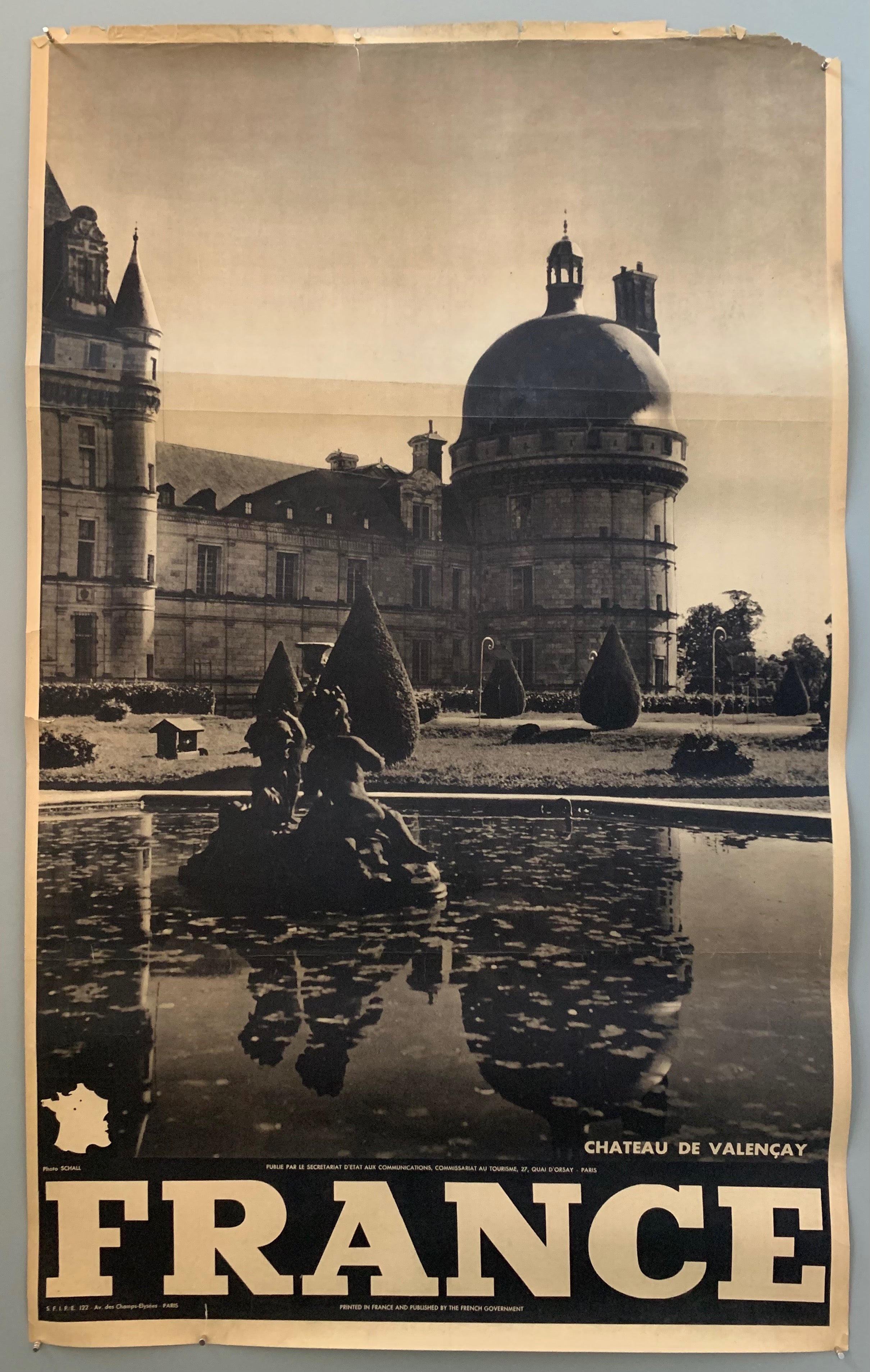 This poster is sepia toned with bold black-and-white writing at the bottom, as well as a tiny map of France. The photograph takes up the majority of the page.The photograph mostly contains a part of the castle that reflects onto a lilypad covered pond. The gardens with shrubbery are between the pond and the domed chateau, and in the middle of the pond is a cherub fountain.