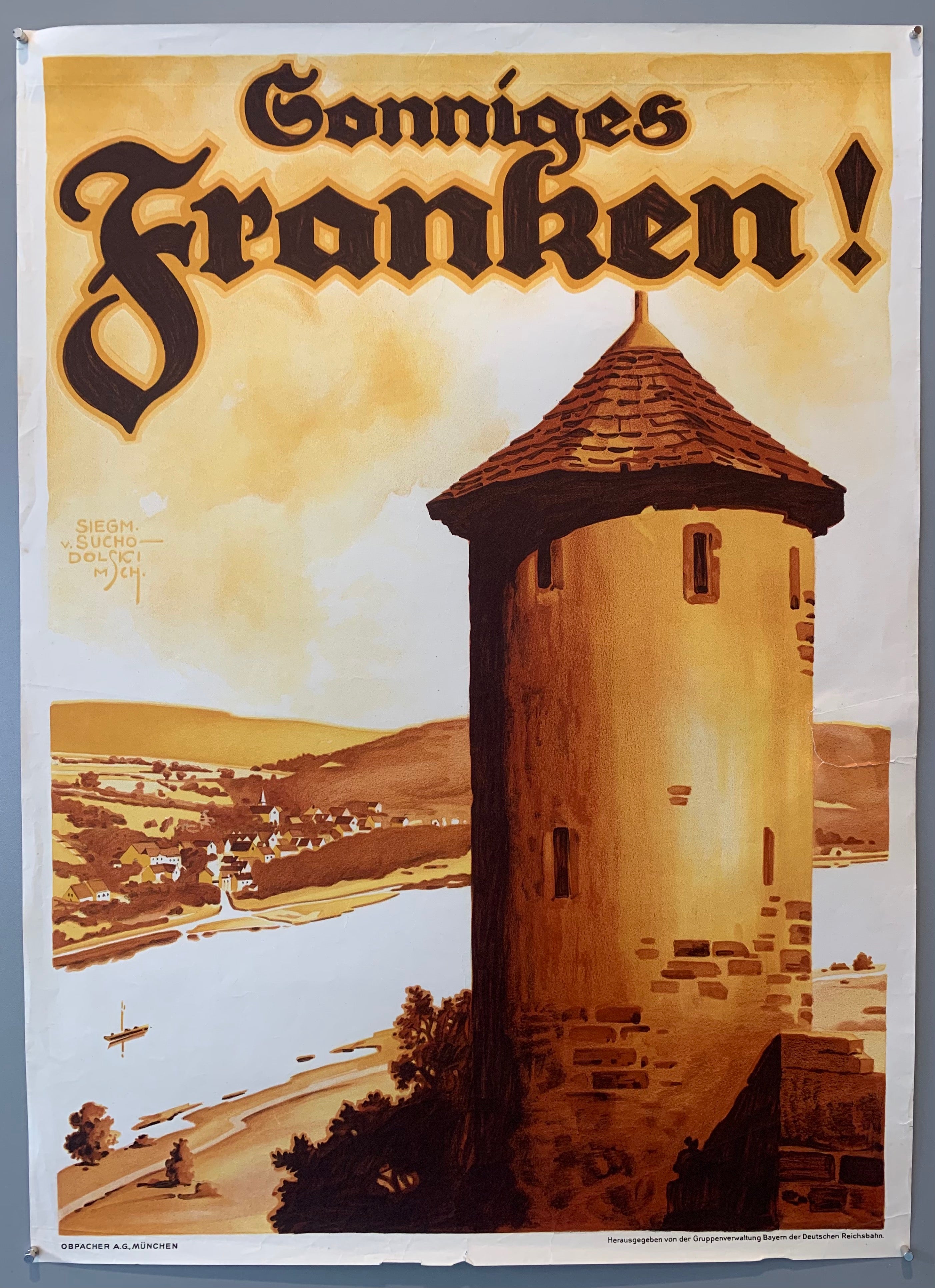 Siegmund von Suchodolski was a German painter, architect and commercial artist. He made many posters for advertisements and travel, amongst children's books and illustrating various publications. This poster shows a Franconian tower overlooking a river with a town in the background. Poster has muted sepia tones.
